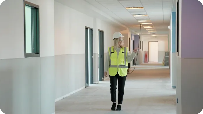 A woman in a safety vest and hard hat walks along a hallway of future Peter J. King classrooma with unfinished walls and exposed flooring. The corridor features several doorways and windows with natural light coming in.