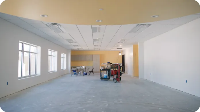 An empty unfinished room showcasing a future Peter J. King classroom, with a yellow ceiling and white walls. There are large windows on one side, and a partially finished kitchenette is visible at the far end. 