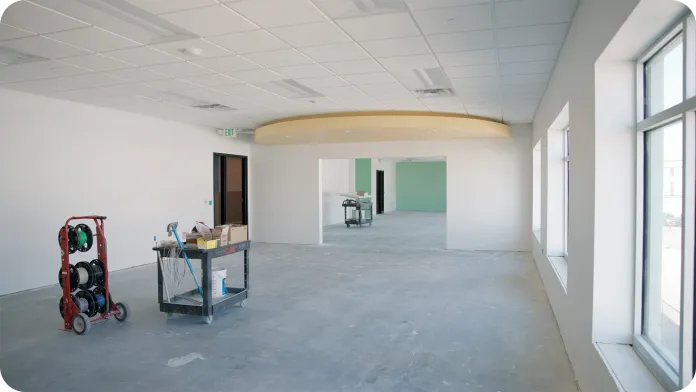 An empty unfinished room showcasing a future Peter J. King classroom, with gray concrete flooring and white walls. Natural light enters through large windows on the right.