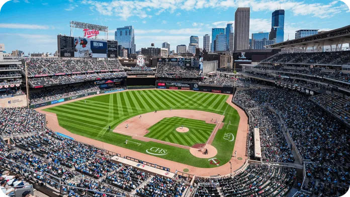 A panoramic view of a baseball stadium filled with spectators for a Minnesota Twins game. The field is well-maintained with green grass, and the skyline of a city with tall buildings is visible in the background under a blue sky with scattered clouds.