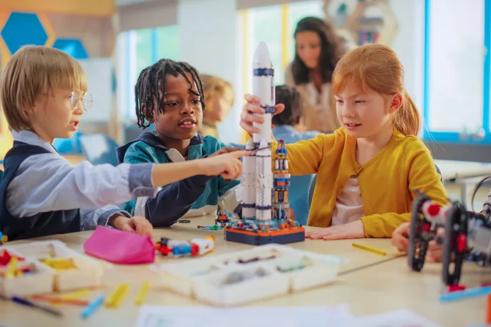 A group of children collaborates at a table to build a model rocket using colorful building blocks. They are focused on their project, with various tools and materials surrounding them, and large windows with daylight in the background.
