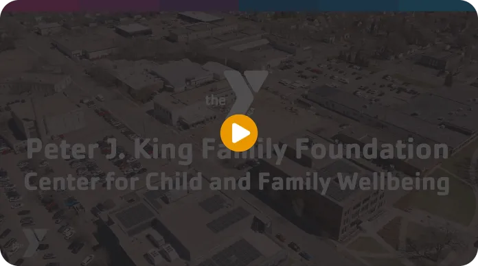 An aerial view of the Peter J. King Family Foundation Center for Child and Family Wellbeing, showing a large building with solar panels on its roof and surrounding parking lots. The image features clear skies and various buildings in the background.