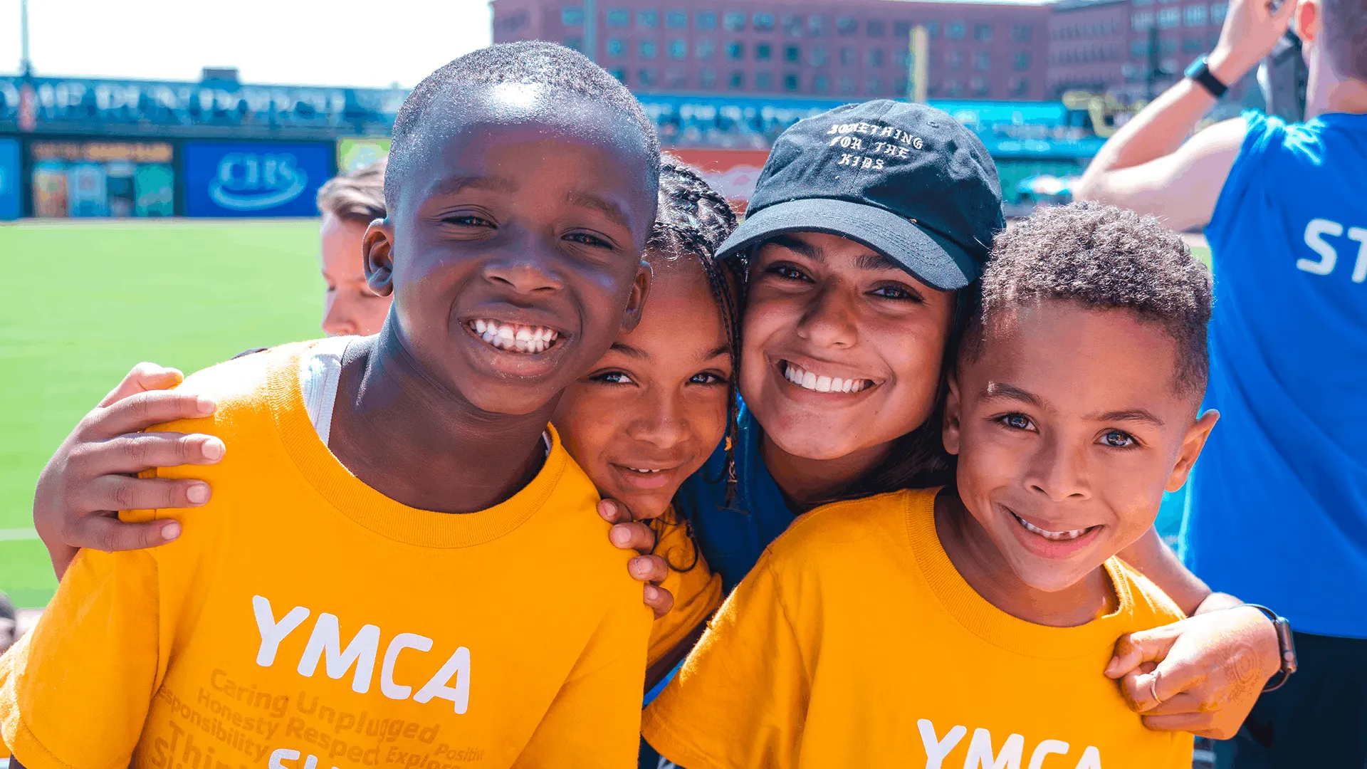 A group of four children and a young woman pose together at a baseball game, smiling and wearing matching yellow YMCA Summer Power t-shirts. The background features a grassy field and a stadium with bleachers.